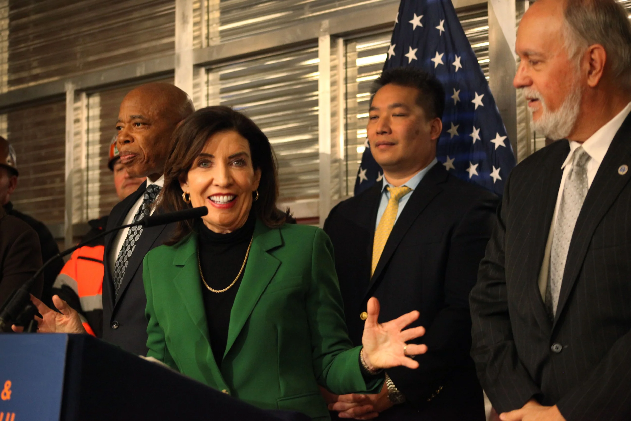 A group of politicians and officials engaged in conversation, including Nancy Pelosi, in a setting with American flags in the background.