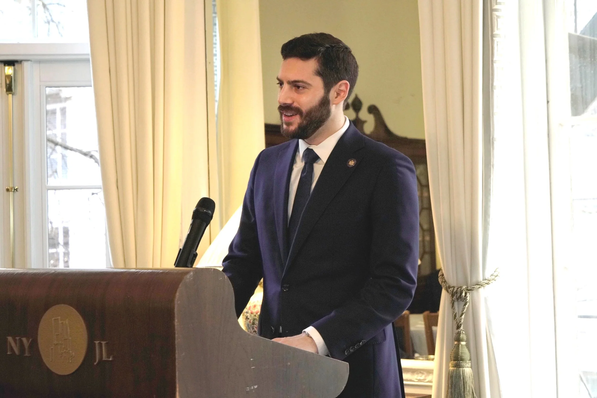 A man with dark hair and a beard wearing a dark suit, white shirt, and blue tie, speaking into a microphone behind a podium in a room with large windows and cream-colored curtains.