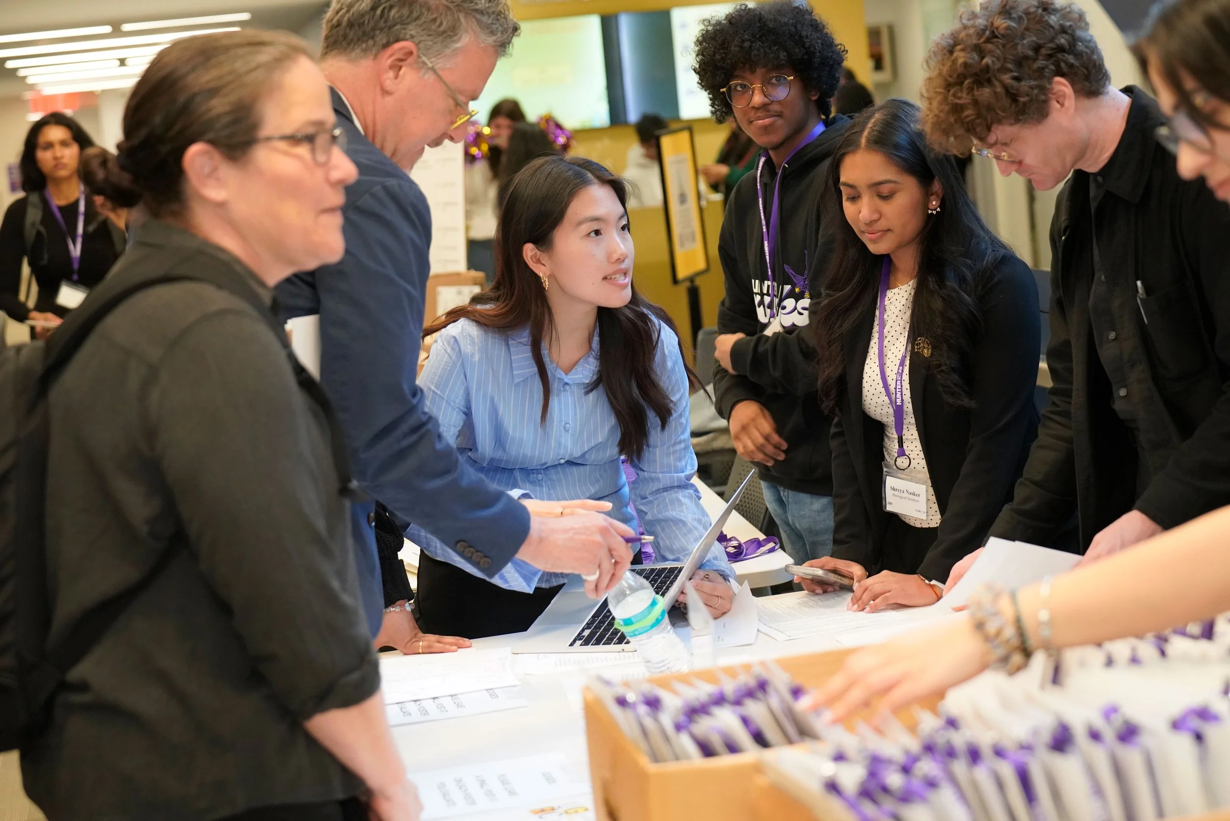 People gathered around a table at a conference or event, engaging in conversation and looking at materials. Some are taking notes or holding papers.