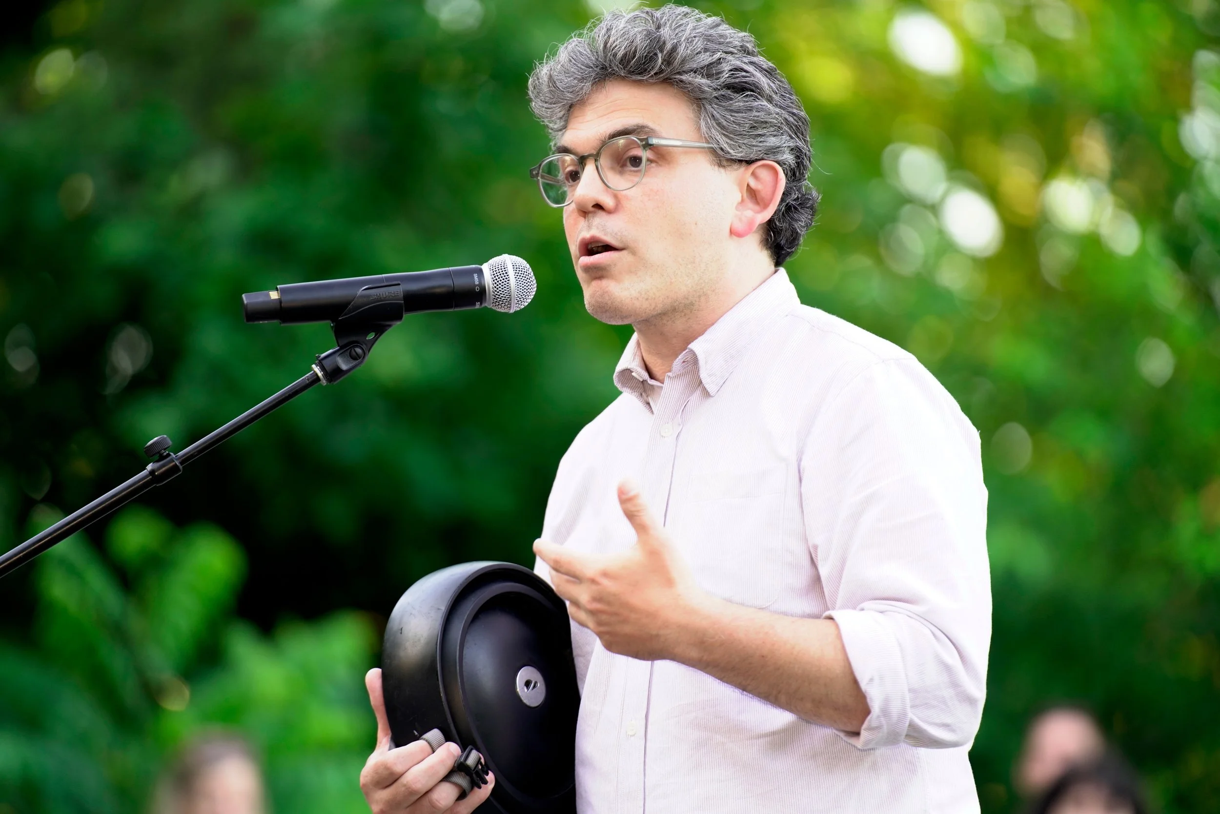 A man with gray hair and glasses speaking into a microphone outdoors surrounded by green foliage.