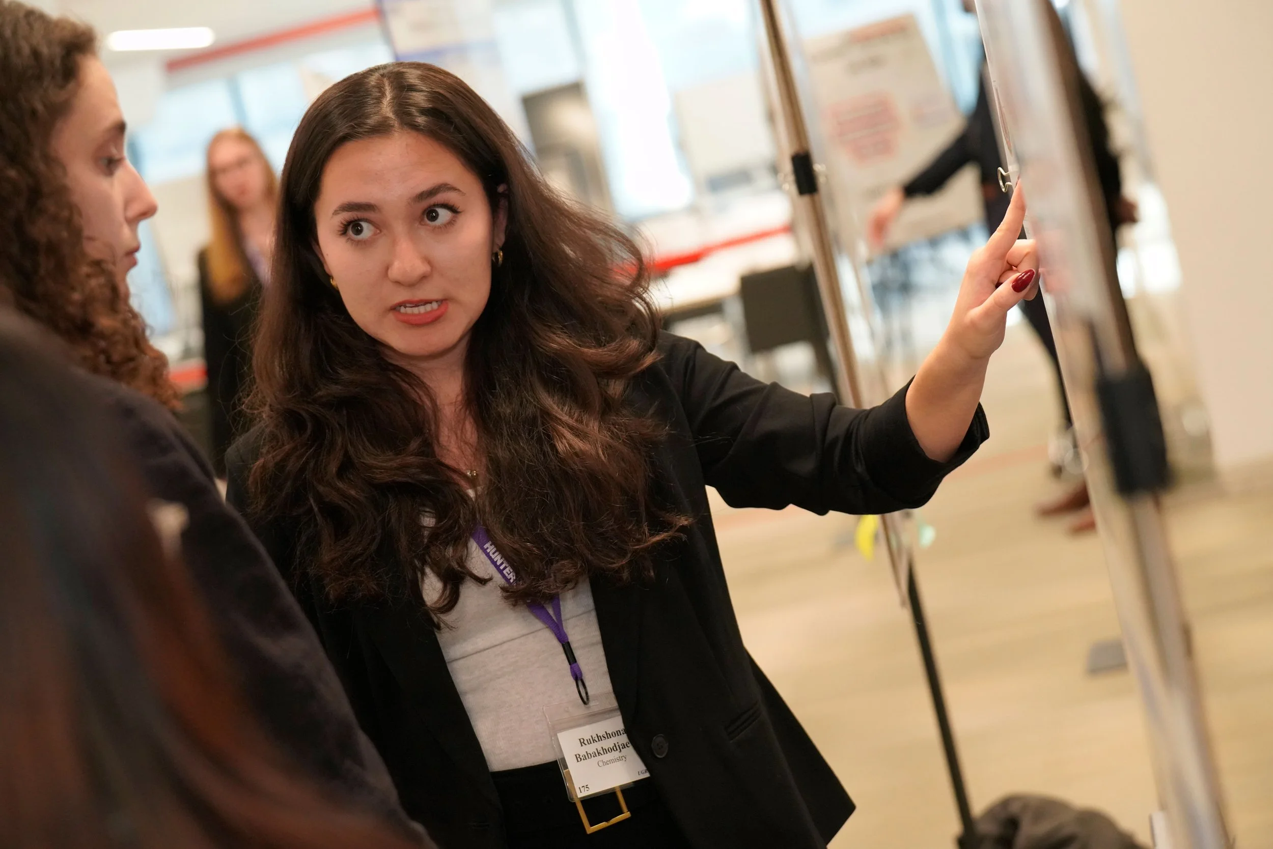 A woman with dark brown wavy hair, wearing a black blazer and a name badge, is pointing at some equipment or display in a room that appears to be a conference or exhibition space. She is engaged in conversation with another person.