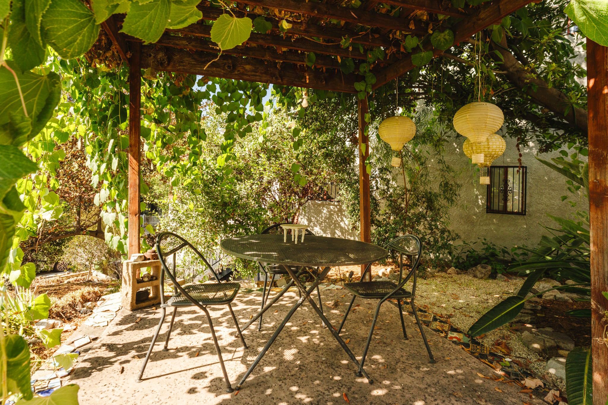 Outdoor dining table beneath a vine-covered wood pergola with hanging lanterns and surrounding garden greenery.