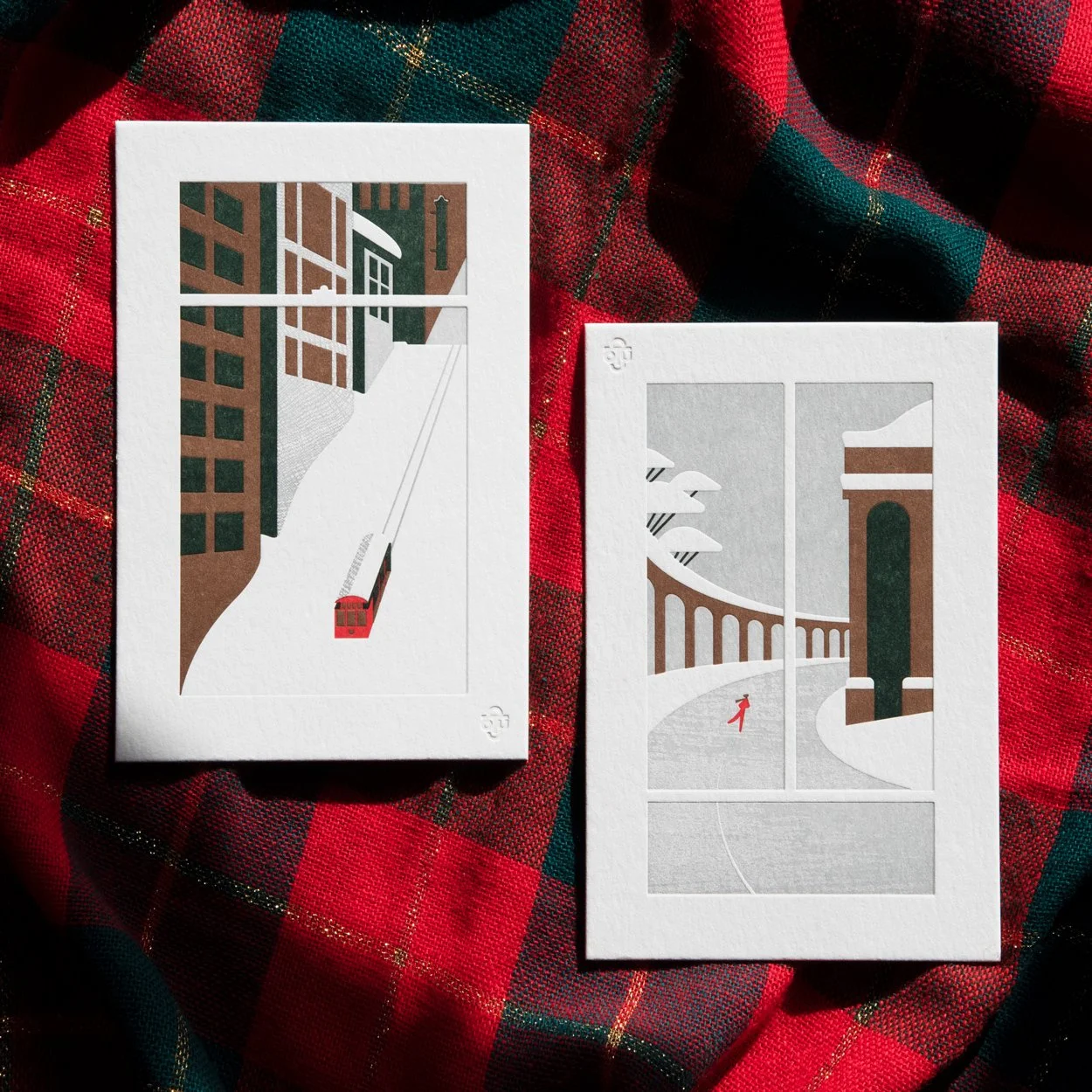 Two illustrated postcards depicting urban winter scenes on a red and green plaid cloth background. The left postcard shows a red streetcar on a snow-covered slope between buildings. The right postcard features a solitary figure walking near a viaduct and an archway in a snowy landscape.