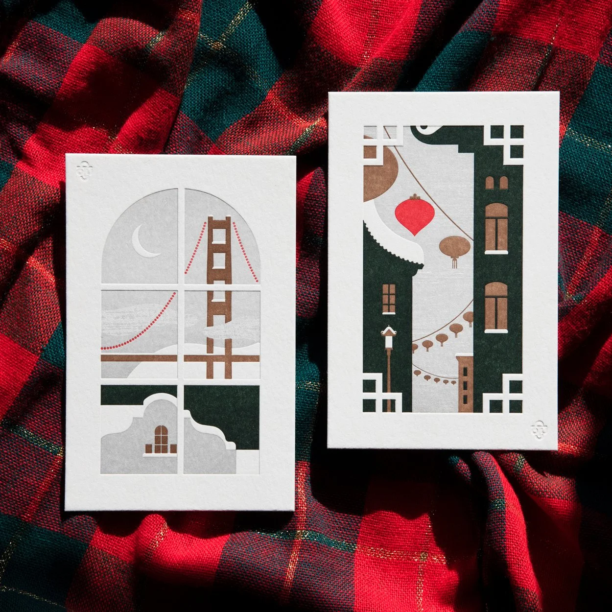 Two minimalist art prints on a red and green plaid fabric. The left print depicts a stylized bridge with a crescent moon, using a grayscale color scheme with red accents. The right print shows an urban setting with traditional lanterns and architectural details, also in a grayscale palette with red accents.