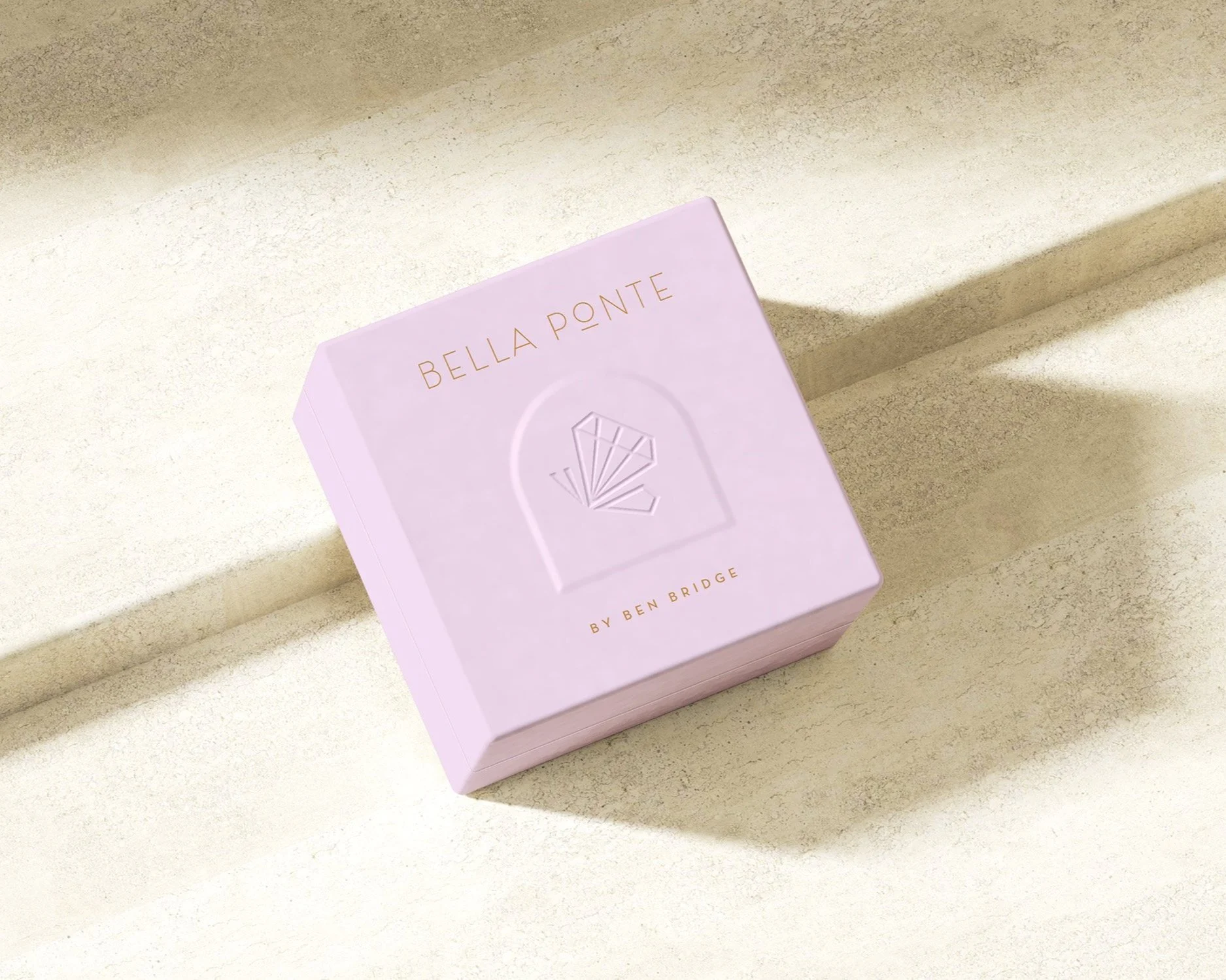 A pink jewelry box on a light stone surface, embossed with a fan design and labeled "Bella Ponte by Ben Bridge."