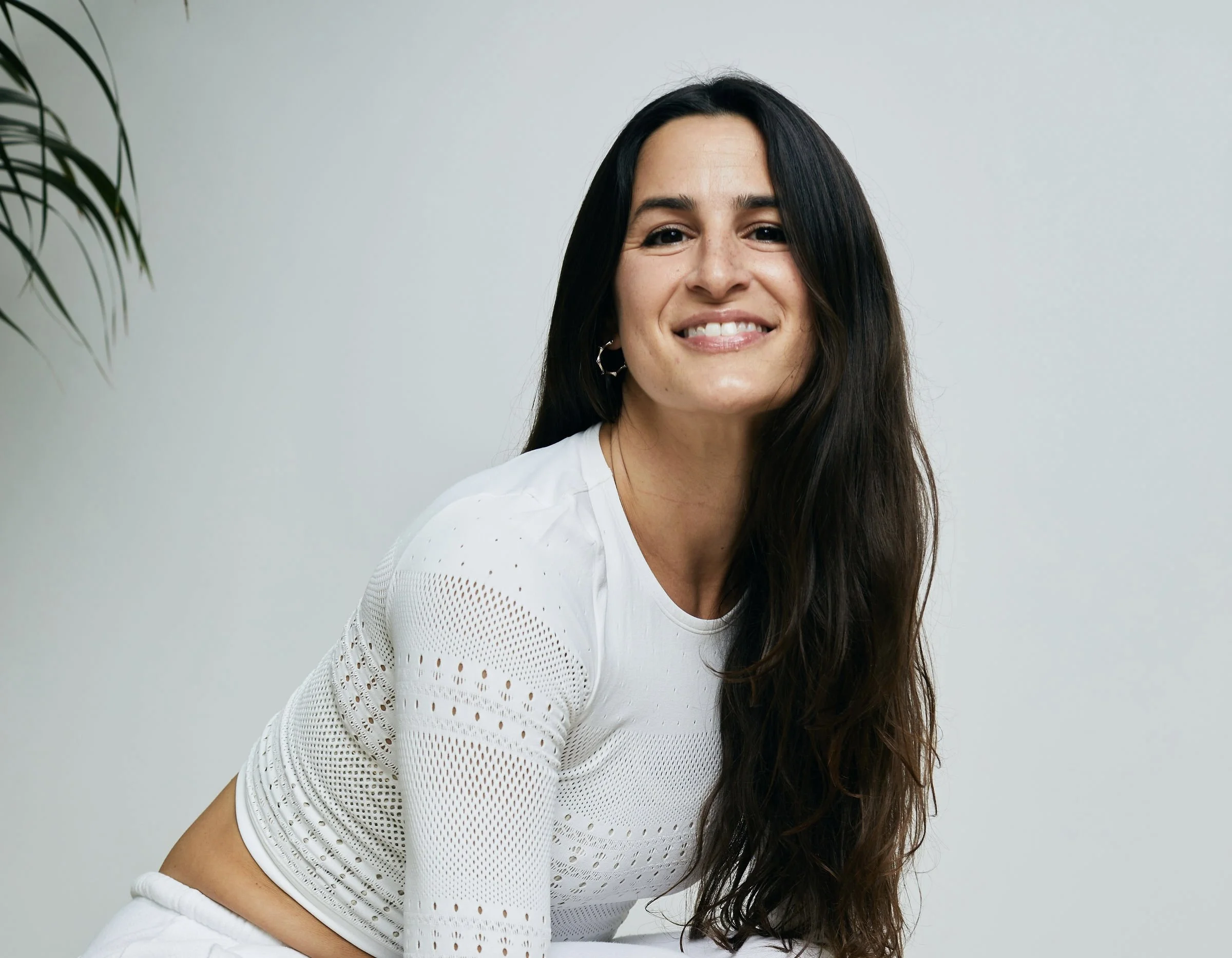 Smiling woman with long dark hair wearing a white textured top, seated indoors beside a leafy plant