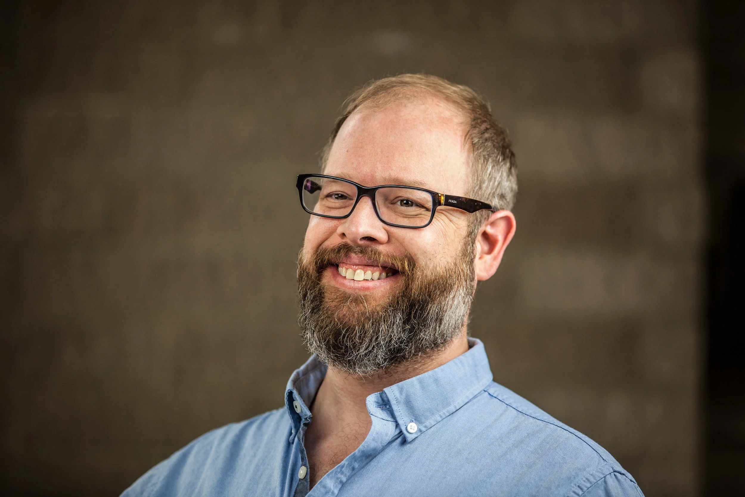 A smiling man with glasses, a beard, wearing a light blue collared shirt, against a blurred brown background.