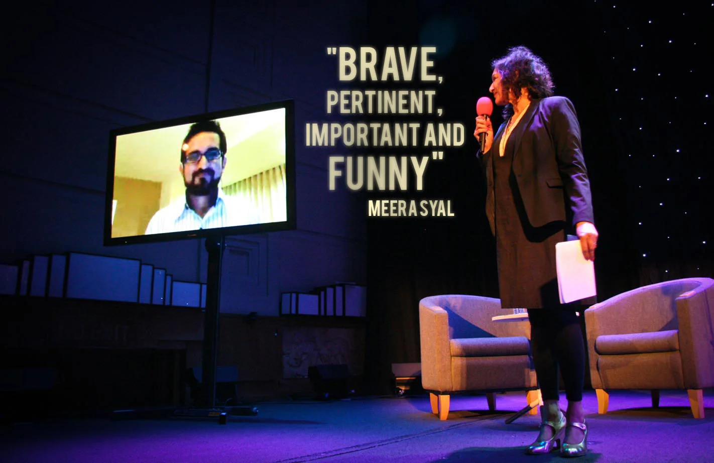A woman with curly hair, wearing a dark blazer and skirt, is standing on a stage speaking into a microphone, with a large screen displaying a man in glasses. The stage has two chairs and a small table, with a quote on the wall reading: 'Brave, Pertin