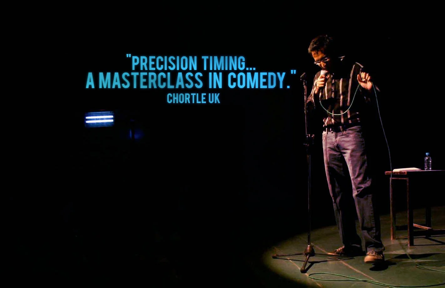 Stand-up comedian on stage with a microphone, a small table with a water bottle and book, and a large screen displaying a quote about comedy.
