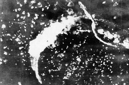 In skies pocked with flak-bursts and waters rippled by bomb blasts, the burning HMS Illustrious is photographed by her assailants as an escort crosses astern.