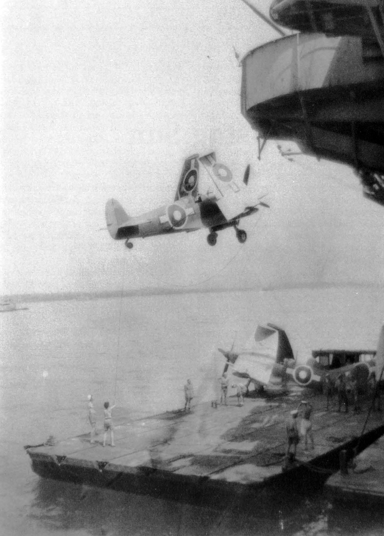A Seafire is slung over the side of what may be HMS UNICORN before being positioned aboard a lighter for transfer in Leyte Harbour.