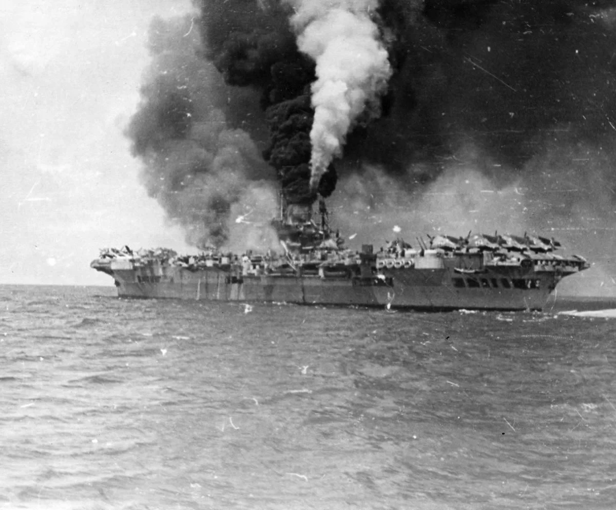 HMS Formidable on fire after the kamikaze strike. The three tones of grey in the smoke rising from the ship tell different stories. The grey is from the burning wreckage of aircraft and fuel on the flight deck. The black oily soot is from Formidable's funnel. The white is steam being vented via emergency systems from the damaged central boiler room.