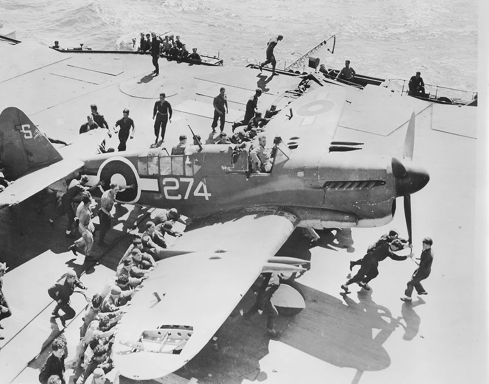 A FireFly from HMS INDEFATIGABLE displays flak damage to its right wing as it is manhandled towards the forward deck park by landing parties.