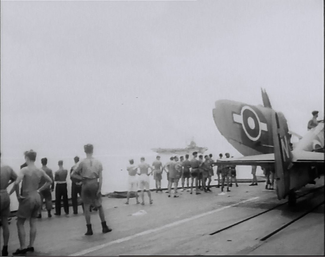 Deck handling crew mill about aboard HMS IMPLACABLE, with a folded FireFly sitting on the dual-track catapult.