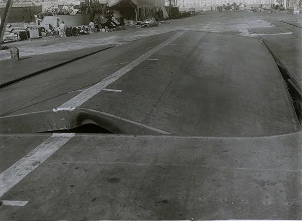 ILLUSTRIOUS's forward lift, buckled upwards by compression from the explosions in the hangar and aft lift well. In the extreme background the aft-lift can be seen sitting upside down on the flight deck. There is no sign of the 'petaled' metal seen in the action photo of the carrier's steaming and smoking flight deck.