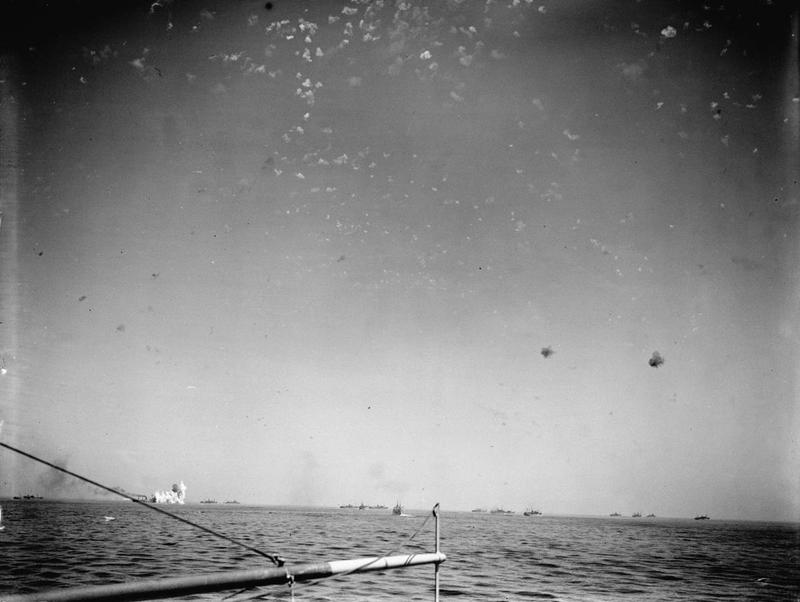 A photo from aboard HMS VICTORIOUS shows a stick of bombs falling near a merchant ship, left, as the sky is filled with flak bursts.