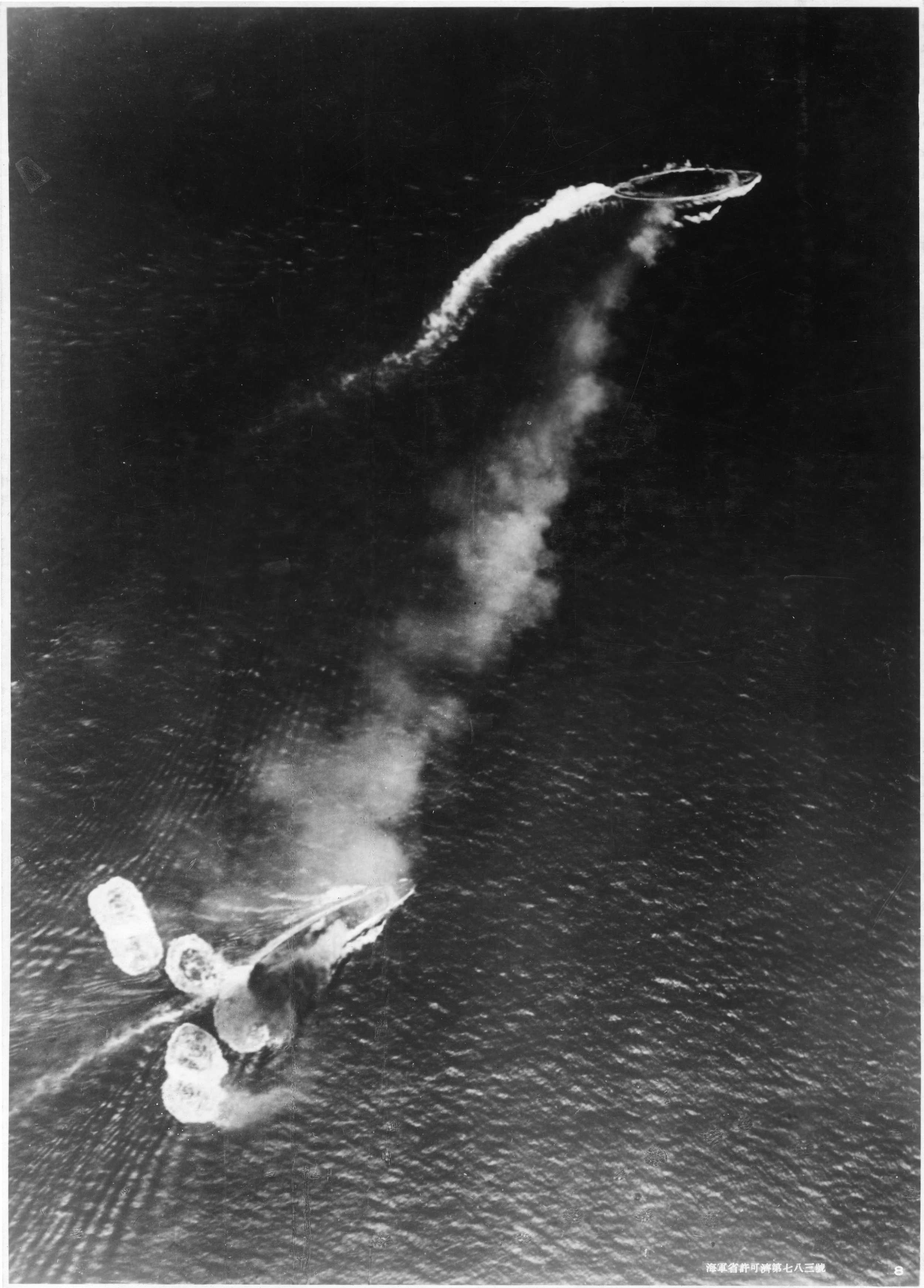 Photograph taken from a Japanese aircraft during the initial high-level bombing attack. Repulse, near the bottom of the view, has just been hit by one bomb and near-missed by several more. Prince of Wales is near the top of the image, generating a considerable amount of smoke. Japanese writing in the lower right states that the photograph was reproduced by authorization of the Navy Ministry.
