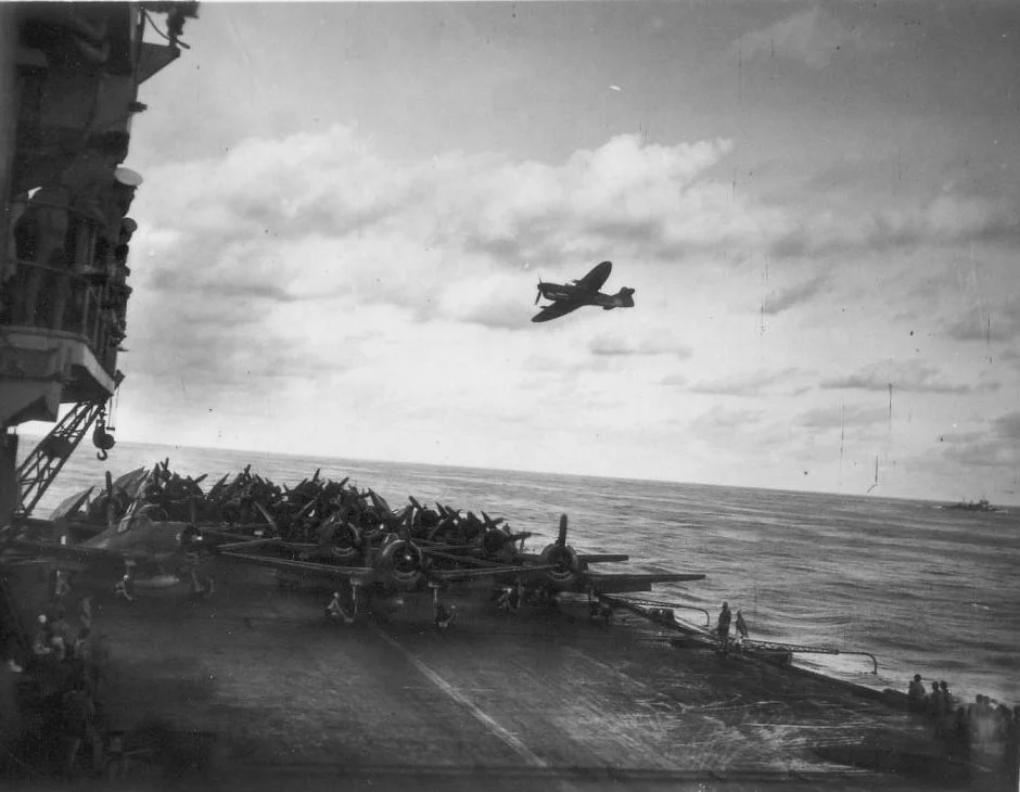 A Firefly from HMS INDEFATIGABLE passes over HMS INDOMITABLE which has a range of Hellcats and Avengers ready for launch.