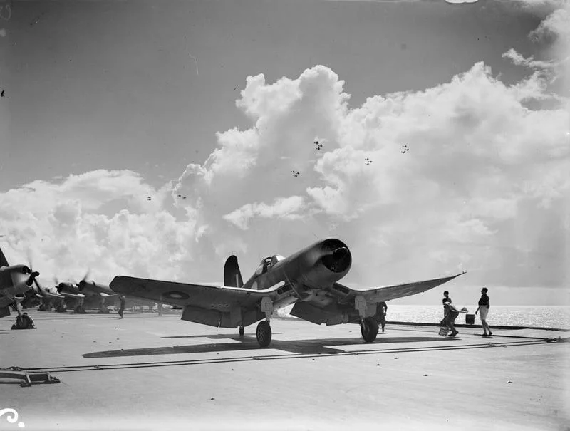 Aircraft are seen silhouetted against the clouds as they 'form up' while a Corsair begins its take-off run from HMS ILLUSTRIOUS.