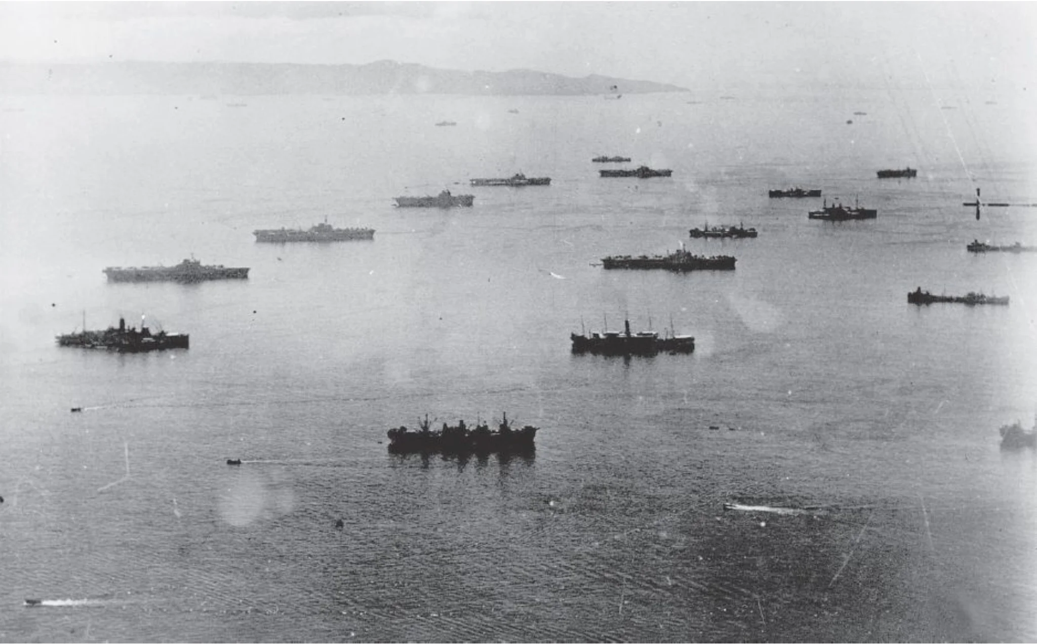 Repair and supply ships are anchored ahead and around the fleet carriers of the British Pacific Fleet. HMS ILLUSTRIOUS (center) is in a row separate from the other carriers as she is being offloaded of useful components before returning to the UK for extended refit and repair.
