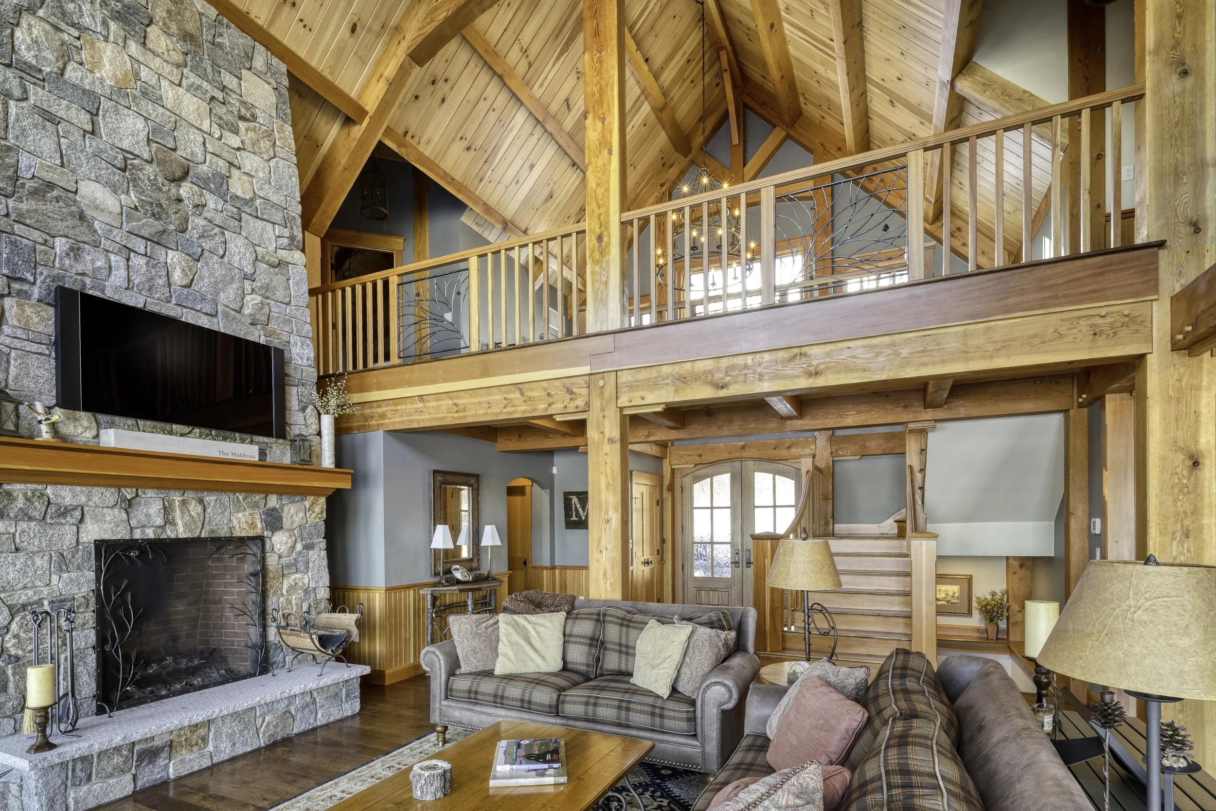 Interior of a cozy wooden cabin living room with a stone fireplace, wooden beams, and a high vaulted ceiling. There are couches, a coffee table, and decorative lights hanging from the ceiling.