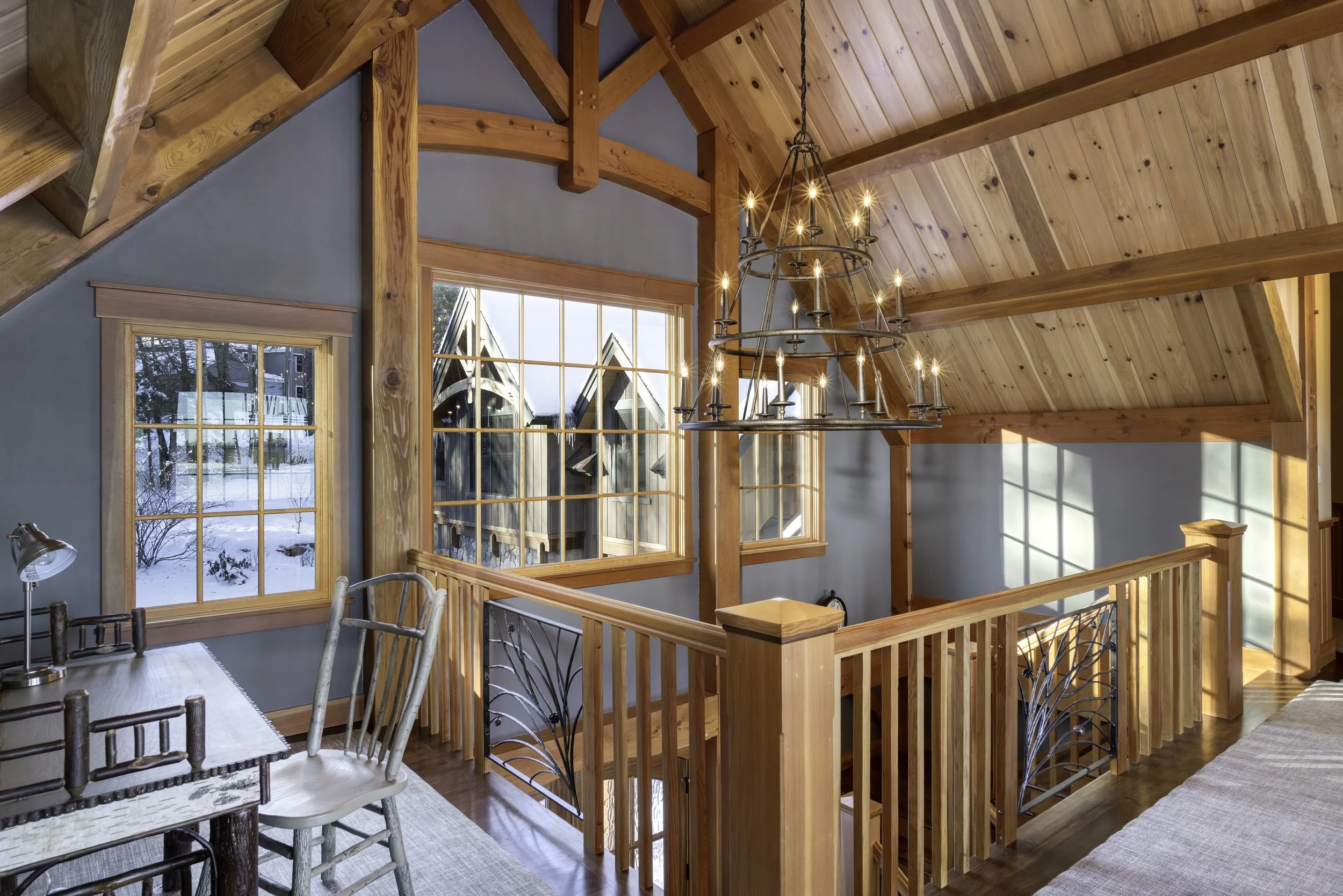 Interior of a wooden house with large windows showing snow outside. A chandelier with candles hangs from the wooden vaulted ceiling. There's a dining table with a lamp and chairs, and a staircase with a railing leading to a lower level.