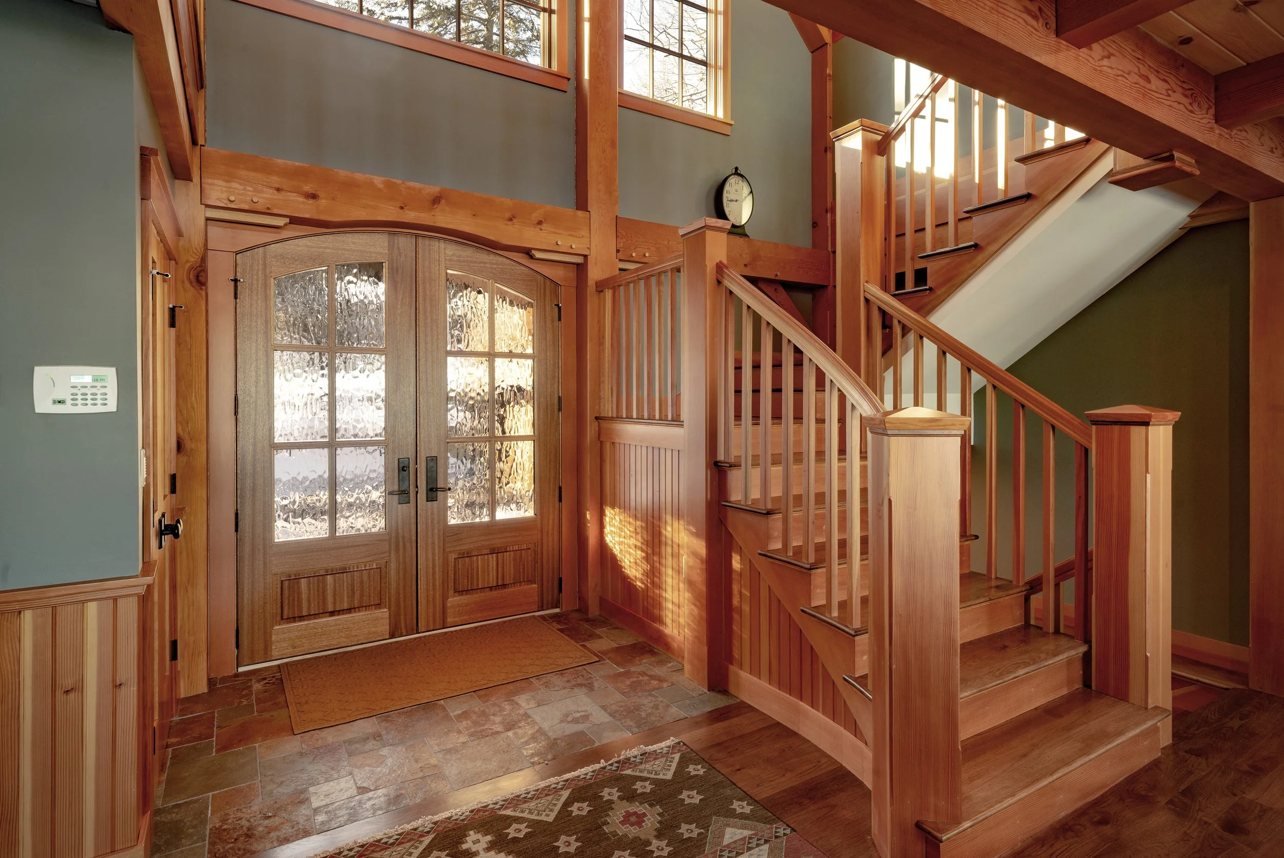 Wooden front door with glass panels, flanked by wooden staircase with railing, large windows above, and a wall clock on a green wall inside a house.