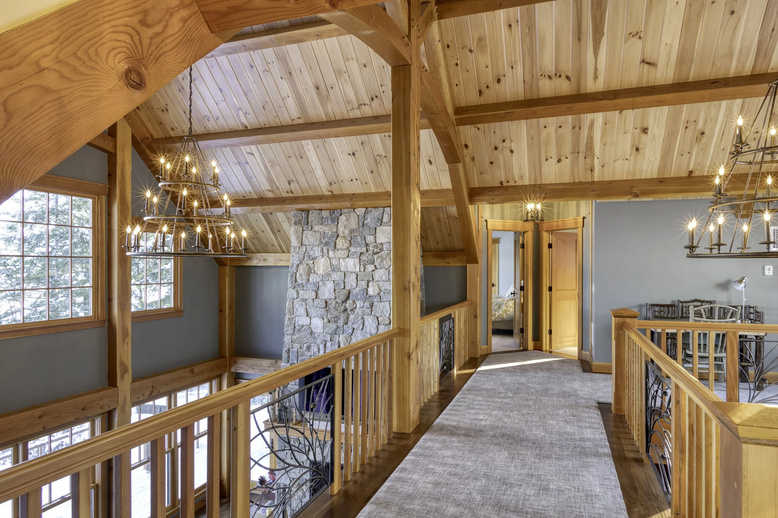 Wooden interior of a house with a sloped ceiling, a stone fireplace, large windows showing trees outside, and chandeliers hanging from the ceiling.