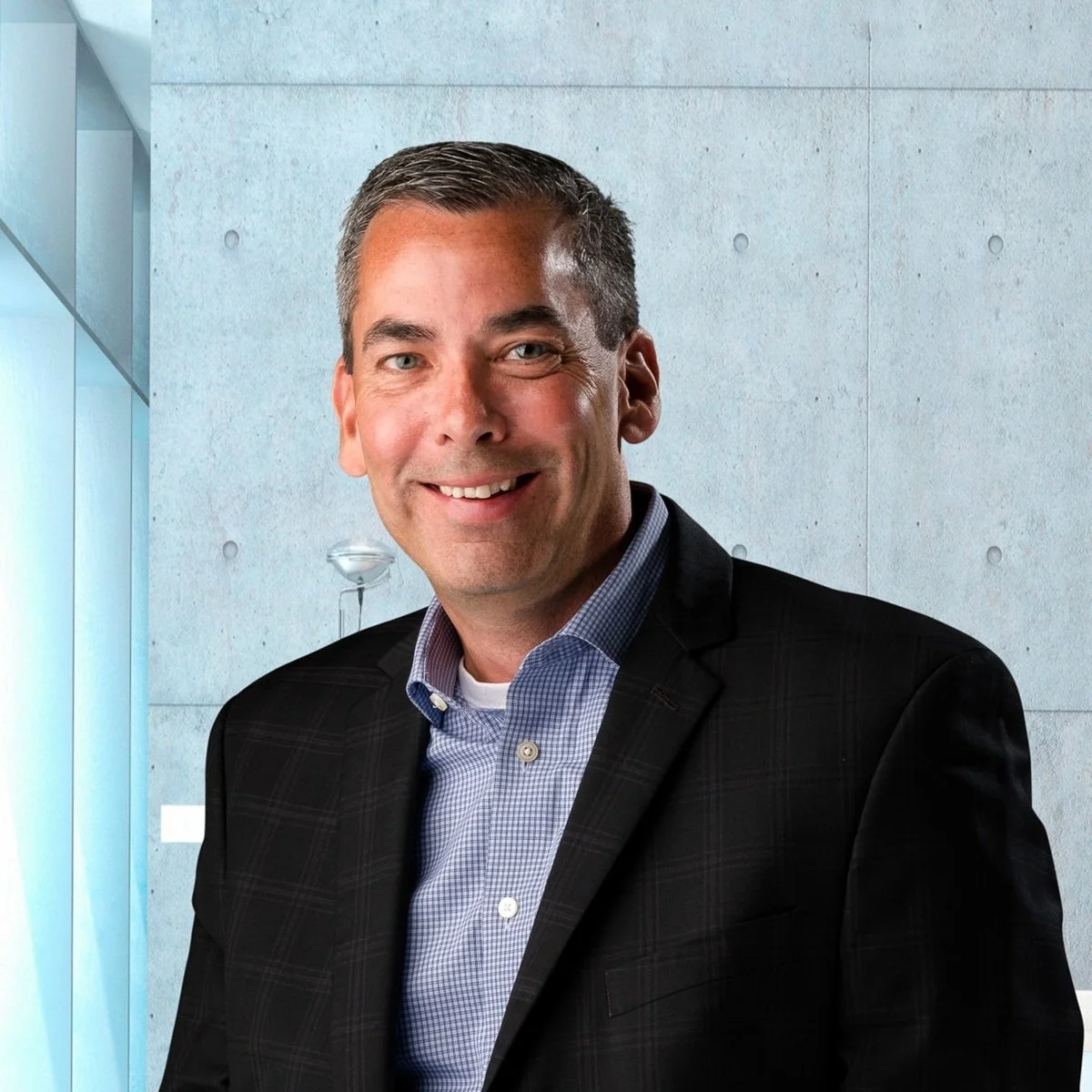 A smiling man with gray hair in a dark blazer and checked shirt pointing towards the camera, standing in a modern building with concrete walls and glass panels.