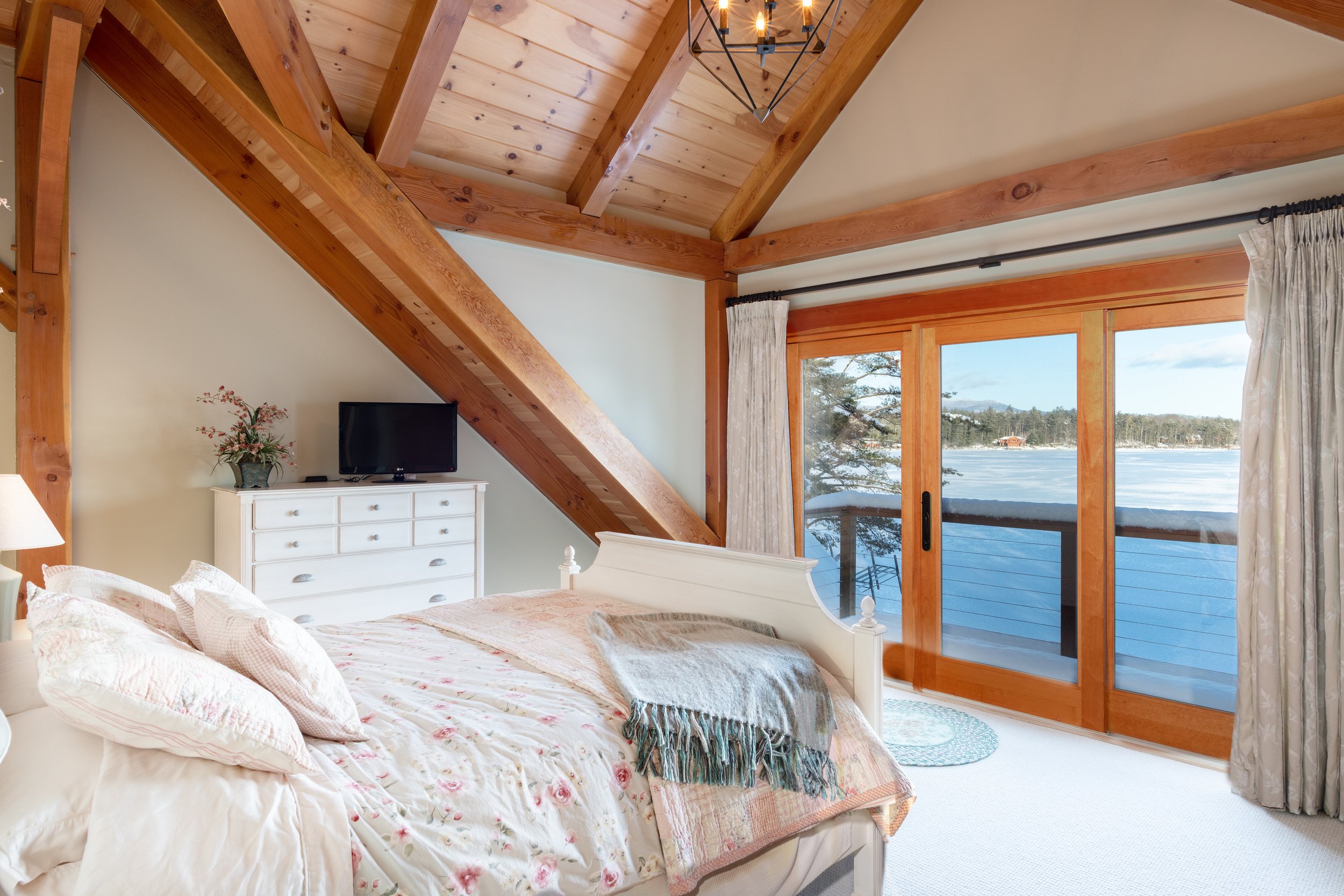 A bedroom with a sloped wooden ceiling, large windows showing a lake view, and a white bed with floral bedding and multiple pillows. There is a white dresser with a small TV and a flower vase, beige curtains, and a chandelier.