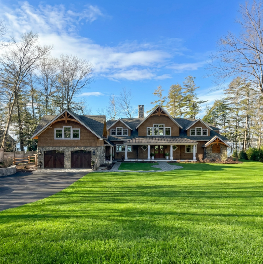 Large house with a well-maintained lawn, stone and wood exterior, front porch, and driveway, surrounded by trees and a blue sky.