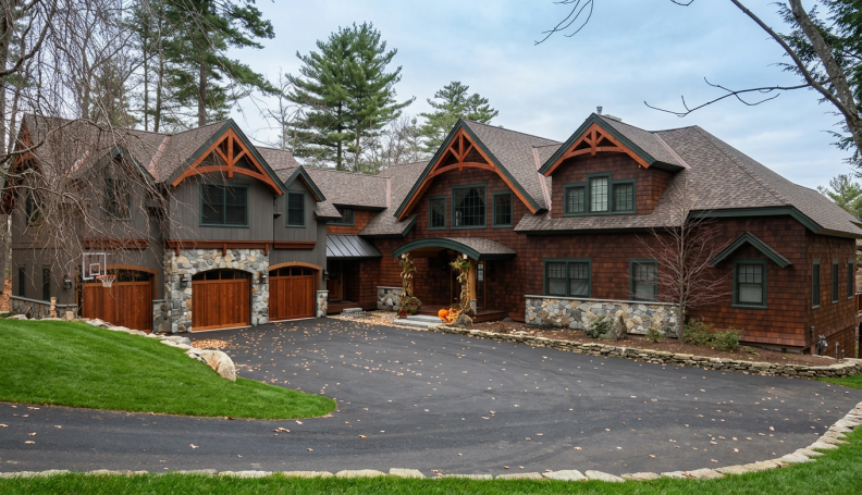 Large house with a stone and wood exterior, multiple gabled roofs, and a paved driveway with fallen leaves, surrounded by trees.