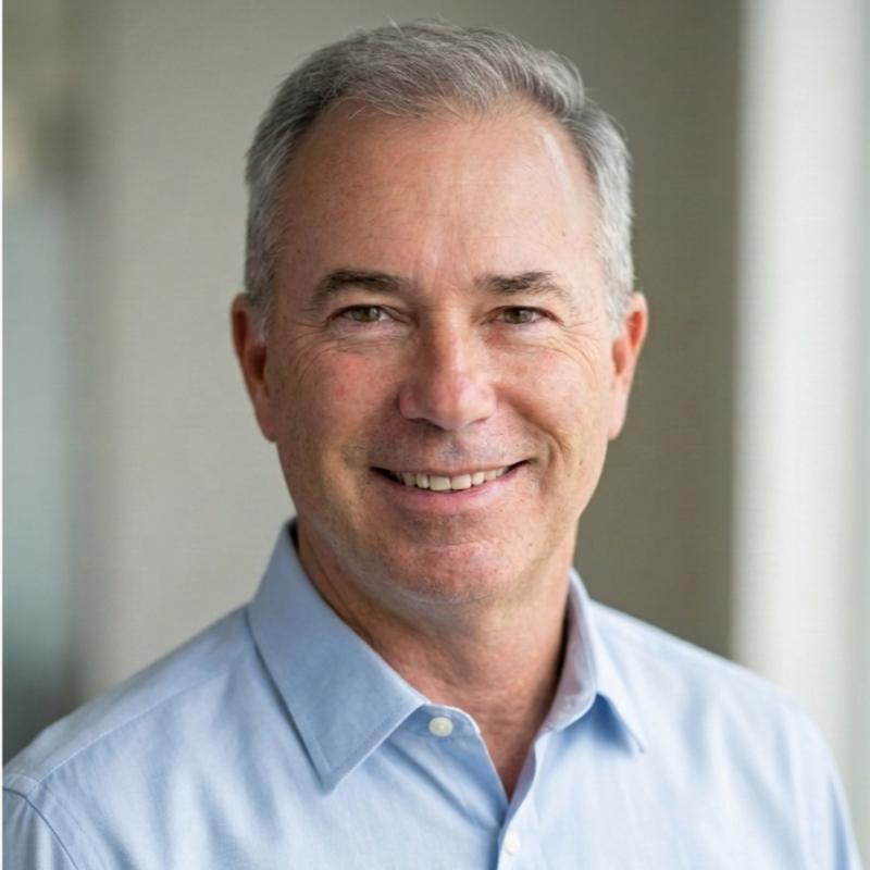 Close-up portrait of a middle-aged man with gray hair, wearing a light blue collared shirt, smiling warmly, in an indoor setting with blurred background.