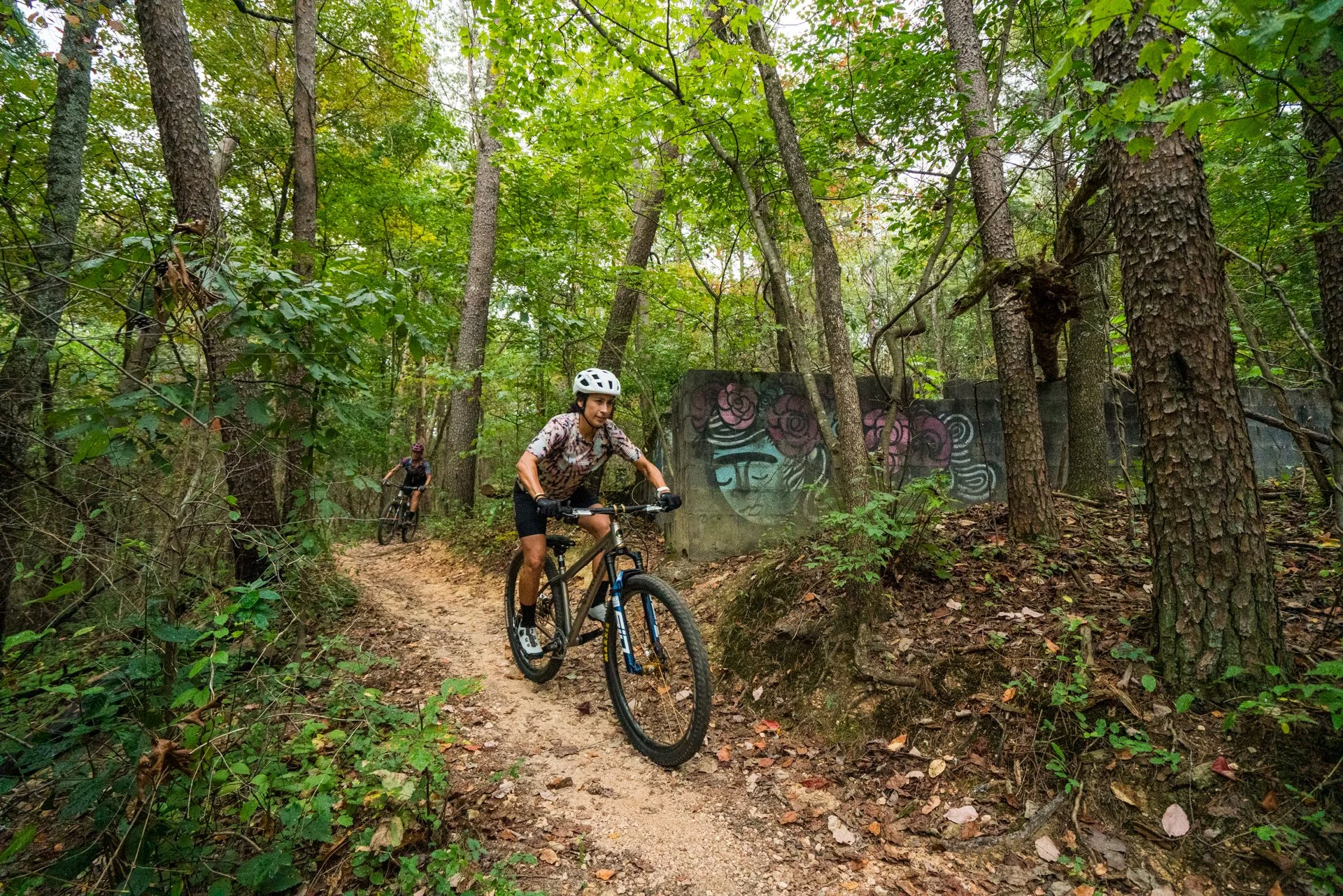 Two women mountain biking on a dirt trail through a green forest. One woman is wearing a white helmet and patterned shirt, the other is in the background wearing a pink helmet.