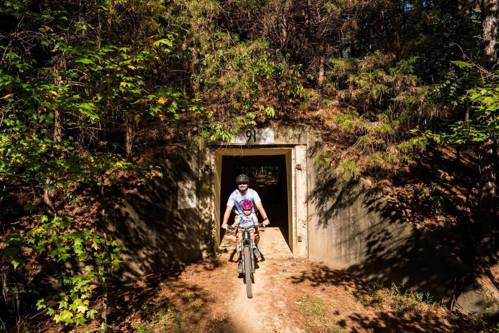 A man and a young girl riding bikes through a tunnel in a wooded area on a sunny day.