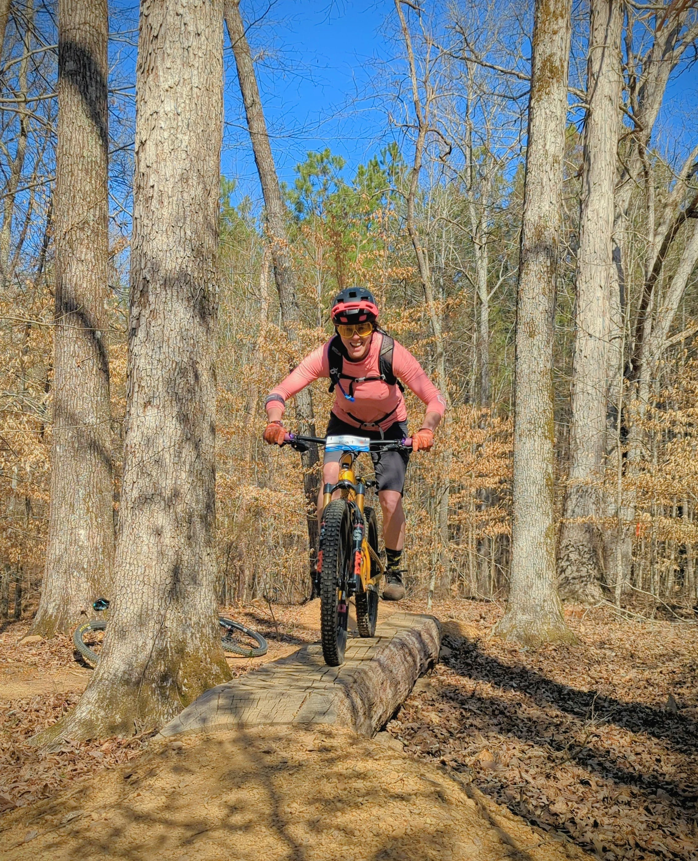 A person riding a mountain bike on a trail through a forest with bare trees and fallen leaves, wearing a helmet and sunglasses, and smiling.