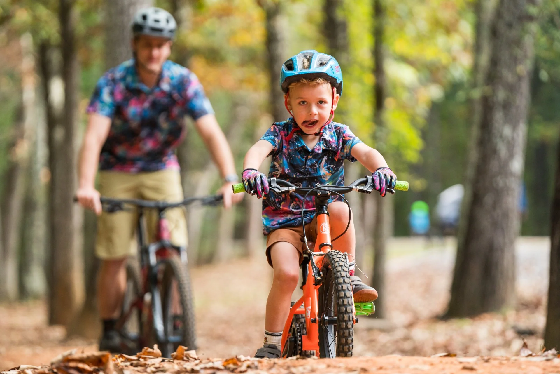 A young boy wearing a blue helmet and colorful shirt rides a mountain bike on a trail in the woods, with a woman behind him.