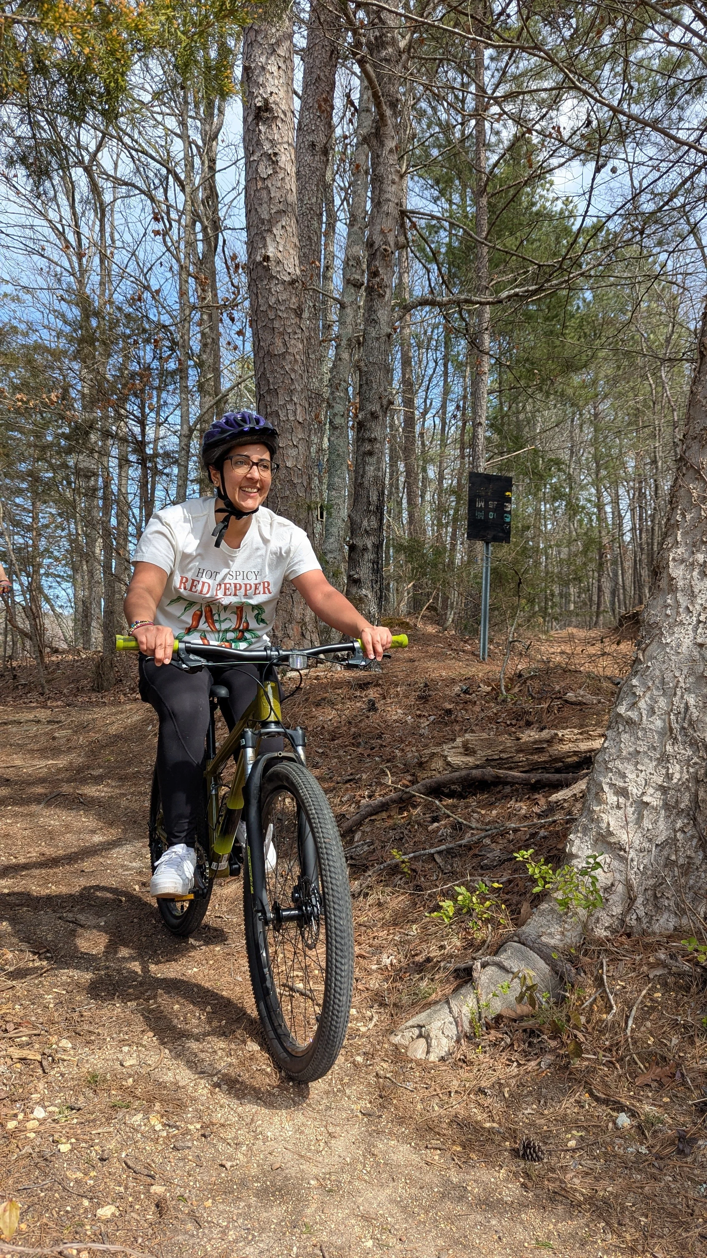 A woman wearing a helmet riding a mountain bike on a dirt trail in a forest with tall trees and some green plants.