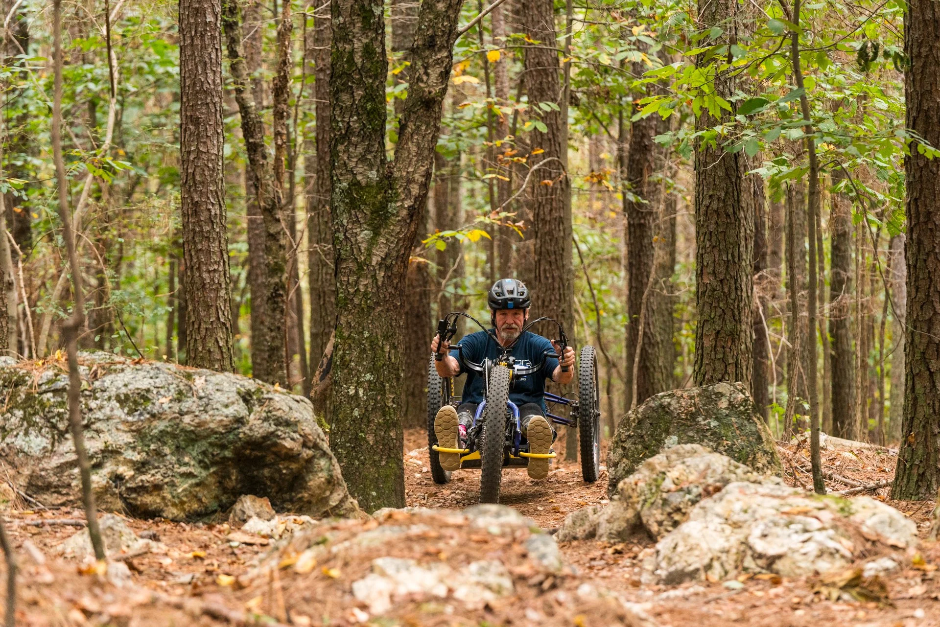 A man in a helmet and gloves riding a handcycle on a rocky forest trail surrounded by trees and rocks.