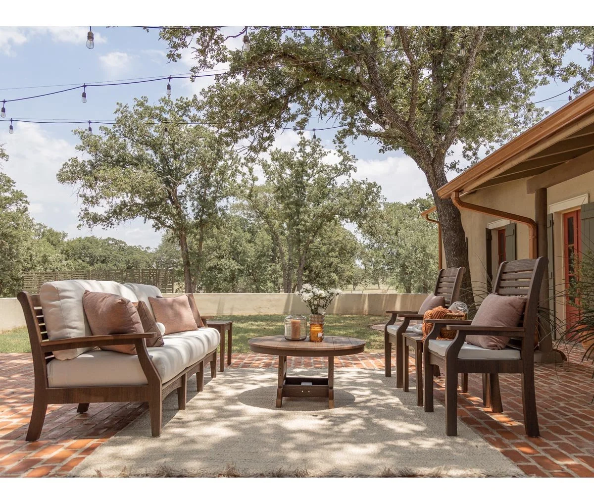 Outdoor patio with poly furniture, including a cushioned sofa and three chairs with pillows, a round coffee table with decorative candles and vases, on a brick patio shaded by trees with string lights overhead.