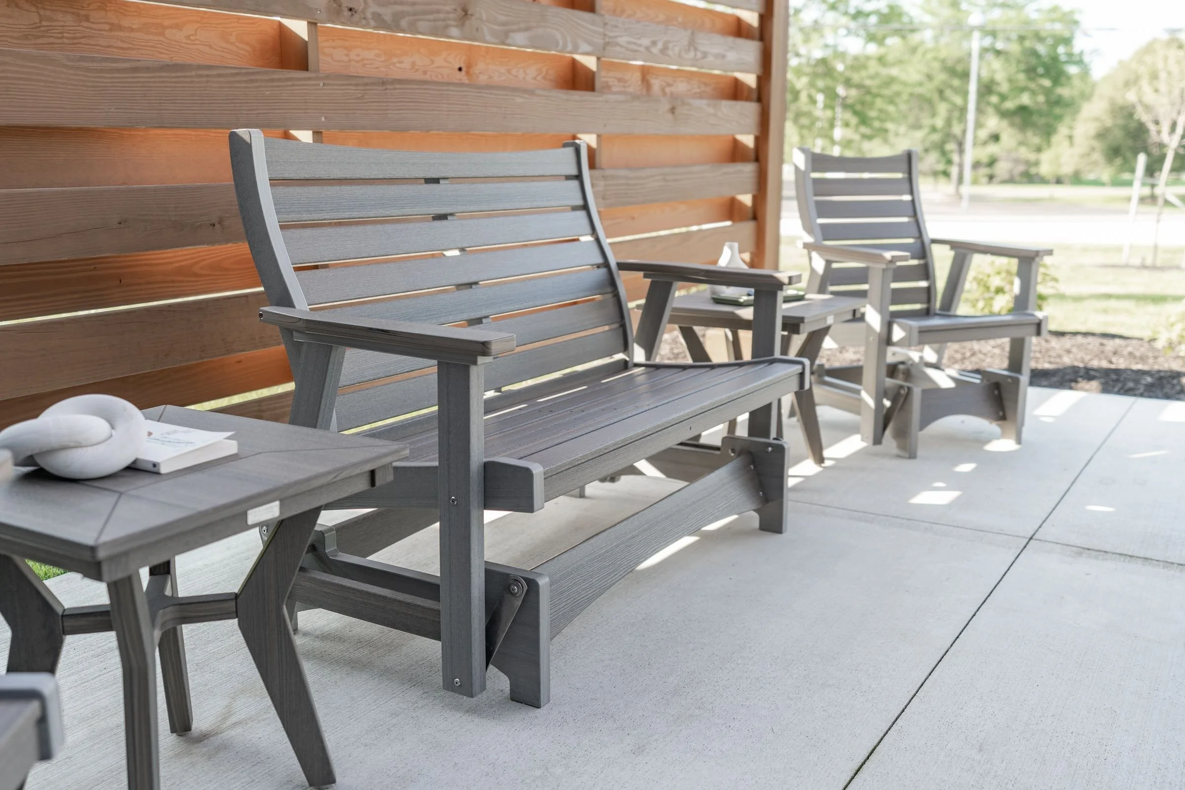 Gray poly glider bench and side tables on a patio, with a modern fence in the background and trees beyond.