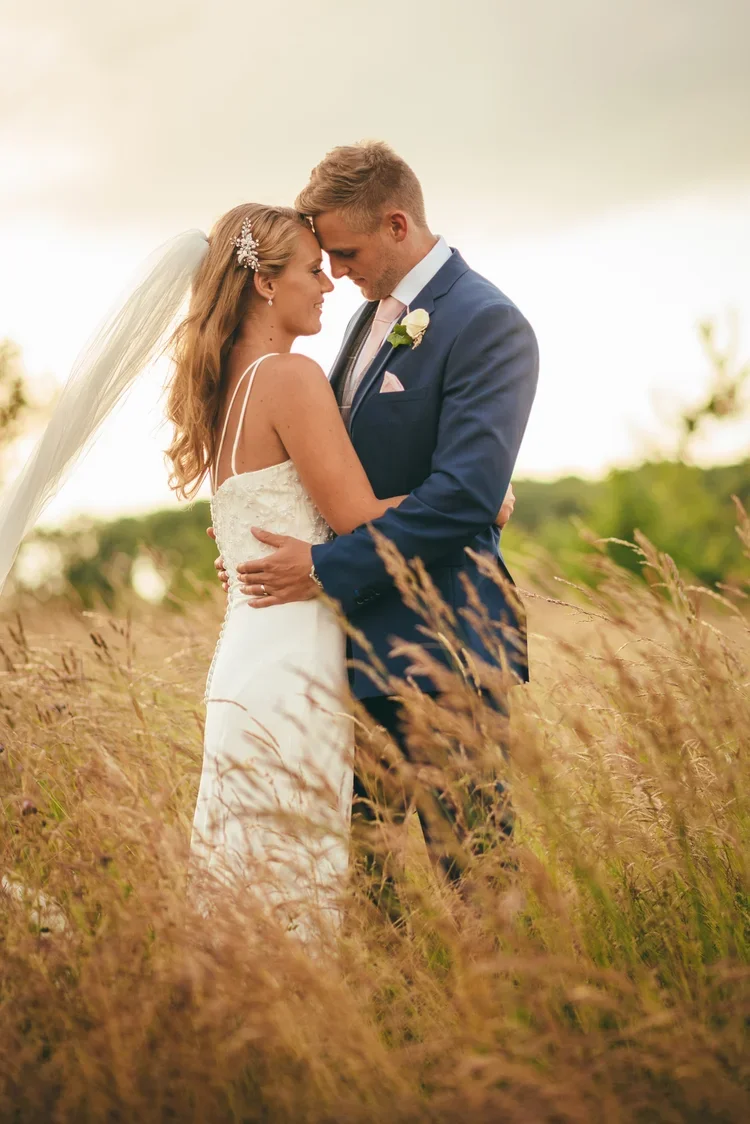 A bride and groom embracing outdoors in a field of tall grass during sunset, with the bride wearing a white wedding dress and veil, and the groom in a blue suit with a pink tie and boutonniere.