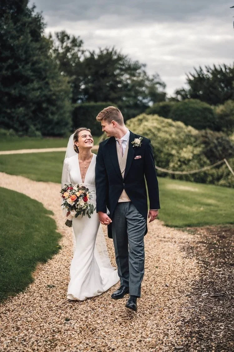 A bride and groom walking hand in hand on a gravel pathway outdoors in a garden setting, smiling at each other during their wedding.