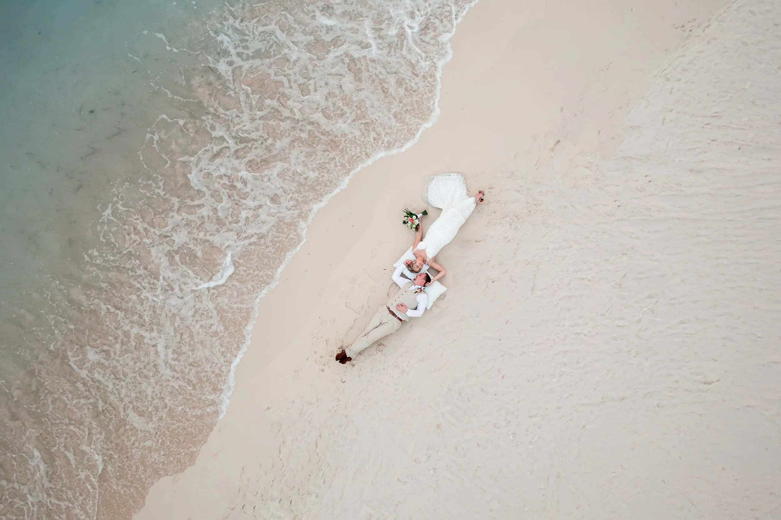  Destination Wedding couple lay on the beach as water approaches for a beautiful drone photo at golden hour 