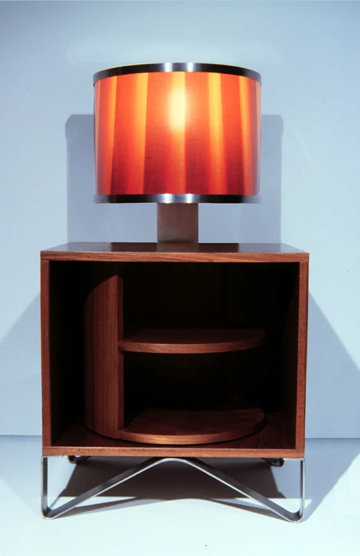   Utsurio Bedside Tables   Teak Veneer + Photographic Film + Aluminum Frame 36”H X 30”W X 30”D  The stripey shade is an enlarged photograph of a traditional fabric lampshade.&nbsp; The cylindrical shelving element can be rotated to hide, reveal or pa