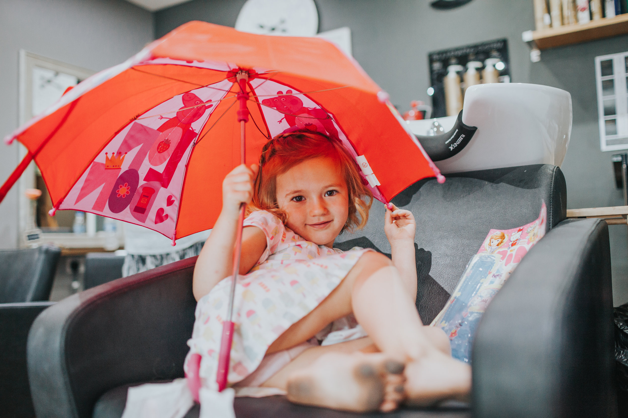  Previously grumpy child, smiles and plays with hair looking into the camera and showing her blackened feet from running around barefoot. She sits under a red umbrella with total rebellious disregard for "It's bad luck to open umbrella's indoors rule