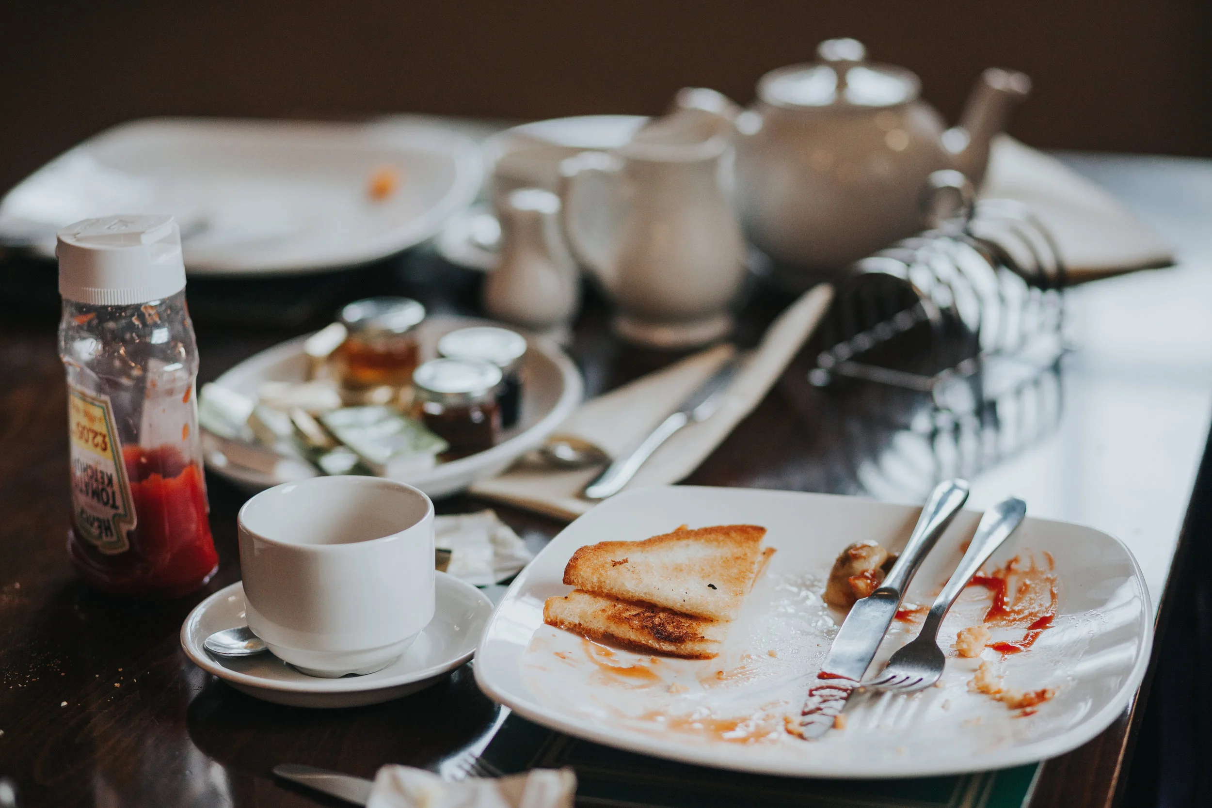Some toast and a bit of ketchup on a plate, an empty coffee cup and some other stuff. 