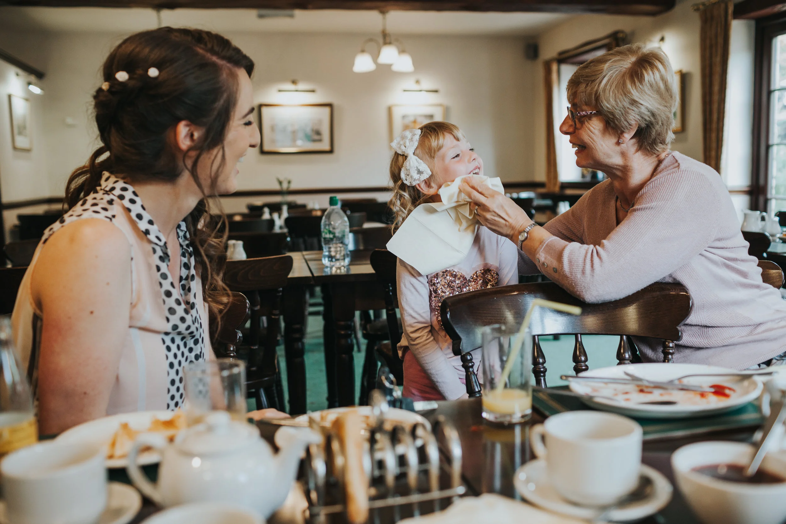  Chipper chid with bow in hair get's ketchup wiped from face by Mother of the Bride, while Bride watches on.&nbsp; 