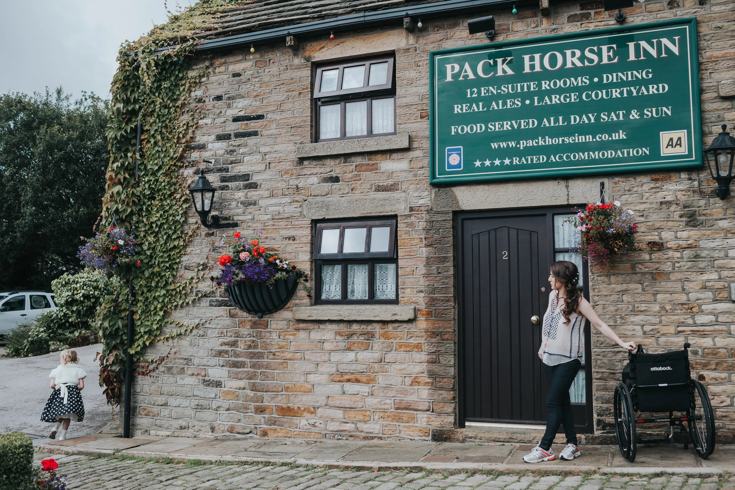 Outside the Pack Horse in, Bride leans casually on Made of Honours Wheel Chair while chipper child pegs it around the corner.