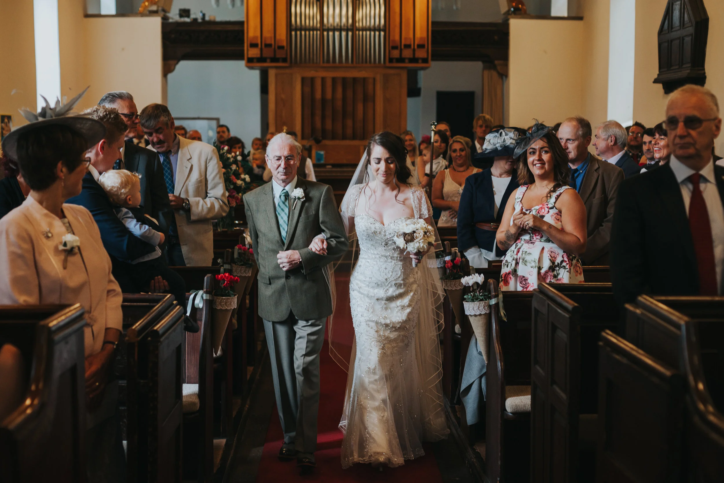 Bride walks down the aisle, caught in window light and close to tears. 
